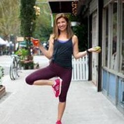 Emily Morgan of the City Skinny in workout attire balances in a yoga tree pose on a city sidewalk, holding a drink in one hand and a piece of fruit in the other, with a tree-lined street and storefront visible behind her.