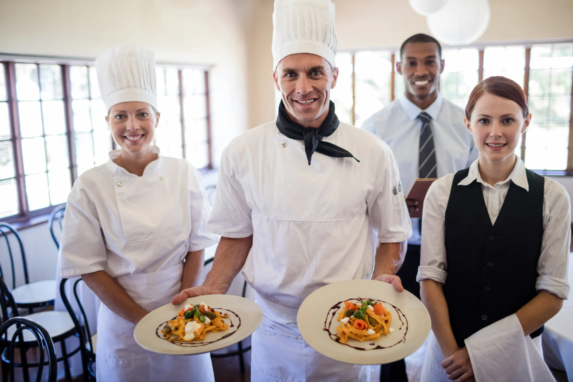 Four hospitality team members in uniform smiling and holding plated dishes in a restaurant dining room