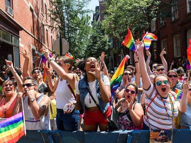 Jubilant crowd of people celebrating at a Pride parade, waving rainbow flags on a sunny city street