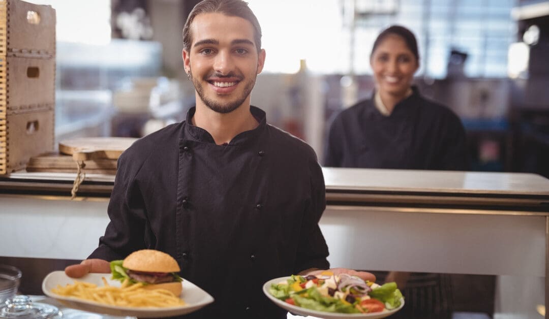 Smiling male server in a black chef coat holding two plates — a burger with fries and a fresh salad — at a restaurant counter