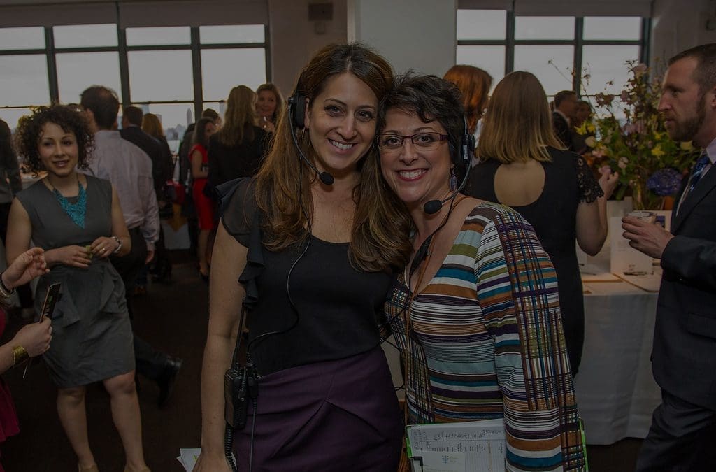 Two smiling women wearing event coordinator headsets posing together at an upscale indoor event