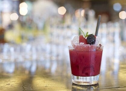 A deep red berry cocktail in a rocks glass filled with crushed ice, garnished with a blackberry, strawberry, and a metal straw, set on a gold bar counter with a softly blurred bar interior in the background.