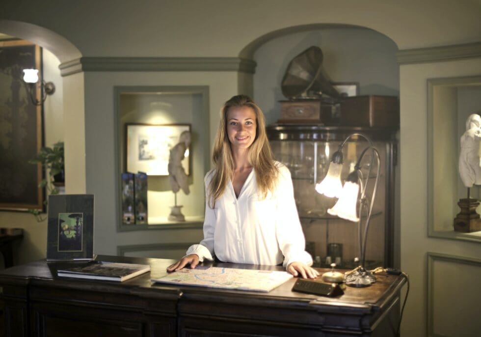 A smiling blonde woman in a white blouse stands behind an elegant hotel front desk adorned with a map, a brass bell, and a desk lamp, surrounded by antique décor including sculptures, a gramophone, and framed artwork in a warmly lit boutique hotel lobby.