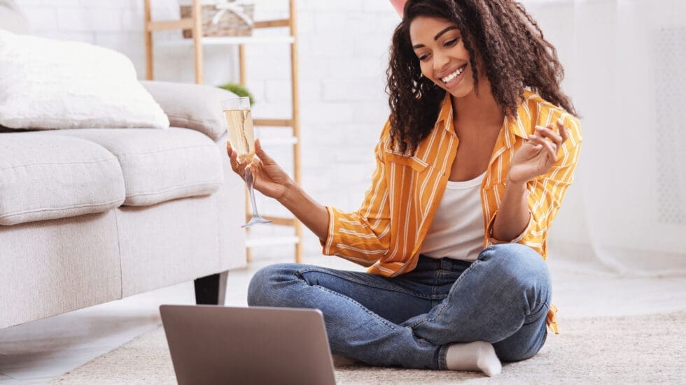 A smiling young woman sitting cross-legged on the floor at home, holding a credit card and gesturing cheerfully while looking at an open laptop in front of her.