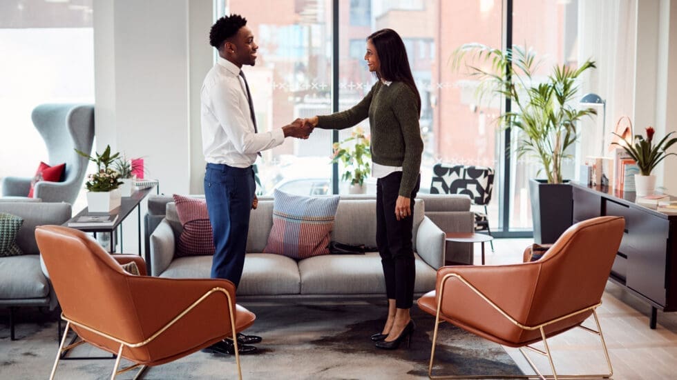A man and a woman in professional attire shake hands and smile in a bright, modern office lobby furnished with grey sofas and orange accent chairs.
