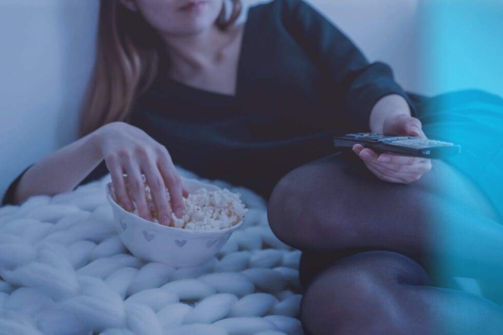 A person relaxing on a couch with a chunky knit blanket, reaching into a bowl of popcorn with one hand and holding a TV remote with the other, bathed in the cool blue glow of a screen.