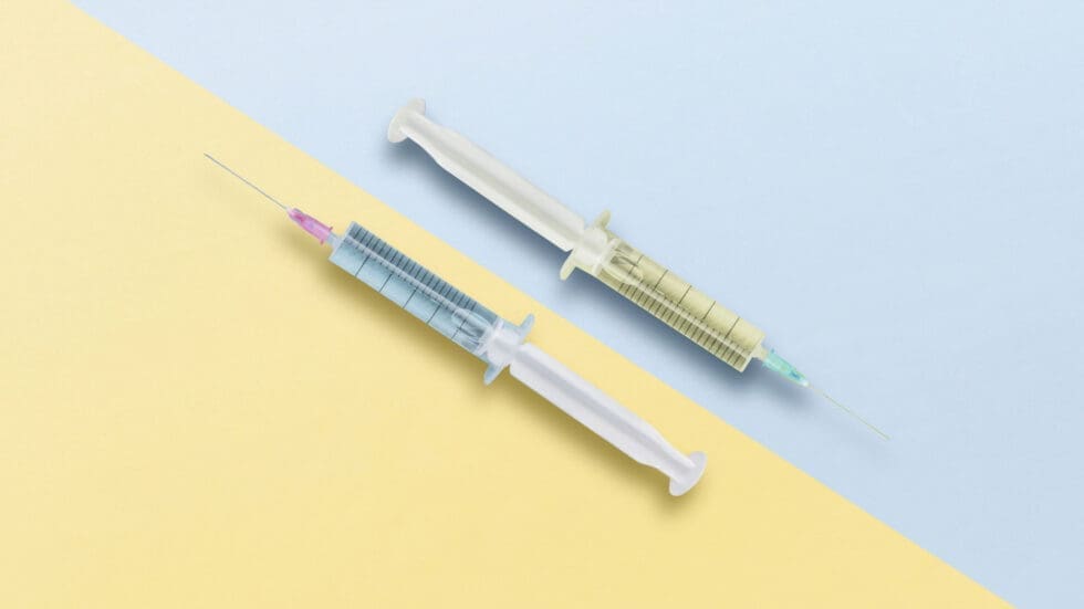 Two medical syringes with capped needles placed diagonally on a split pastel background, one half light blue and the other pale yellow.