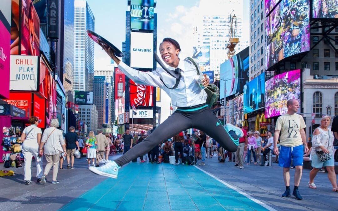 A joyful woman in a white shirt and black leggings leaps mid-air in a wide split jump in the middle of a bustling Times Square, New York City, holding serving trays in both hands, with colorful billboards, crowds of pedestrians, and skyscrapers filling the background.