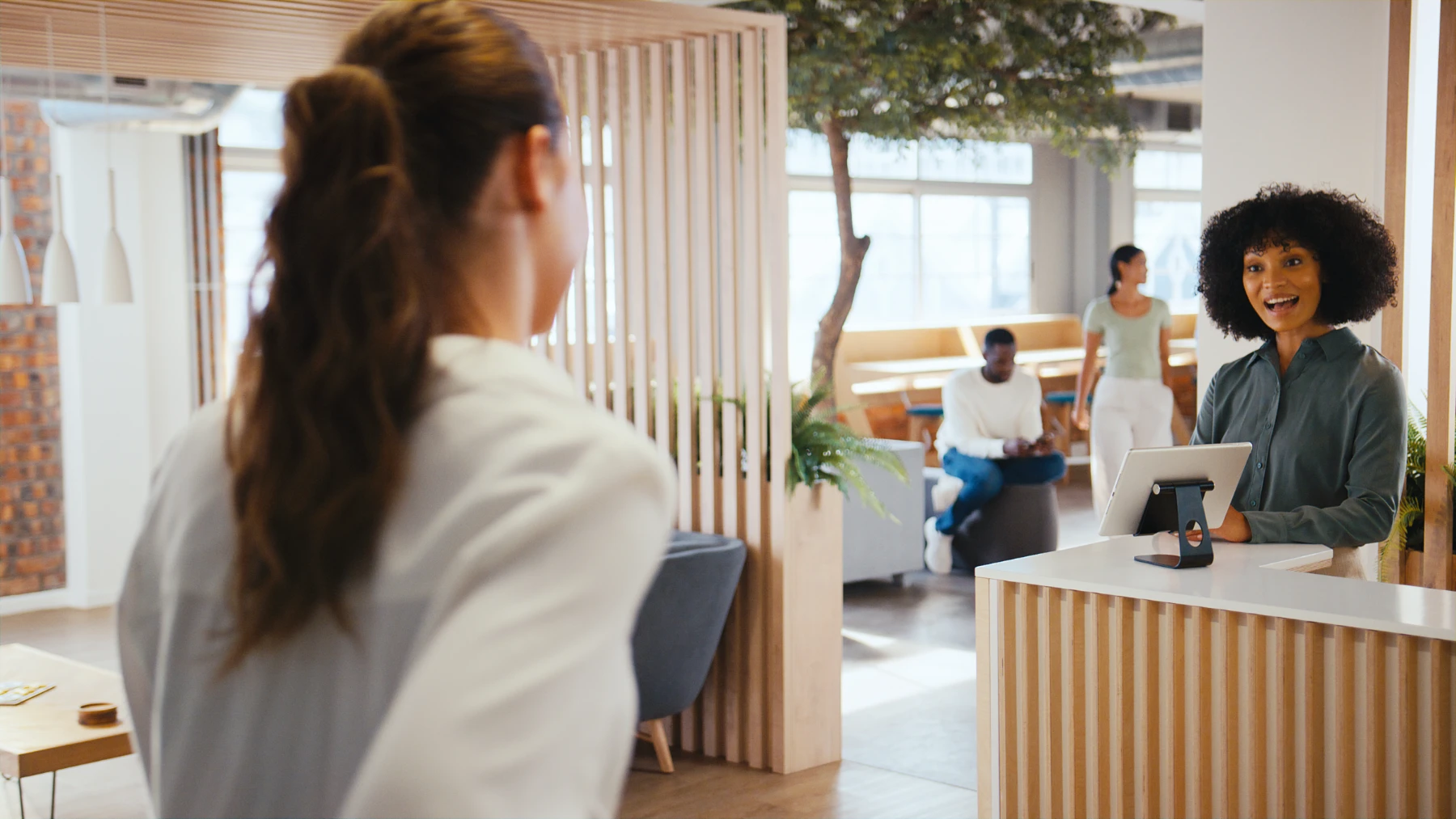 smiling-receptionist-greeting-visitor-in-modern-office-lobby-hmg-plus. A smiling receptionist with natural hair greets a visitor at a bright, modern office lobby with wood accents and a live tree.