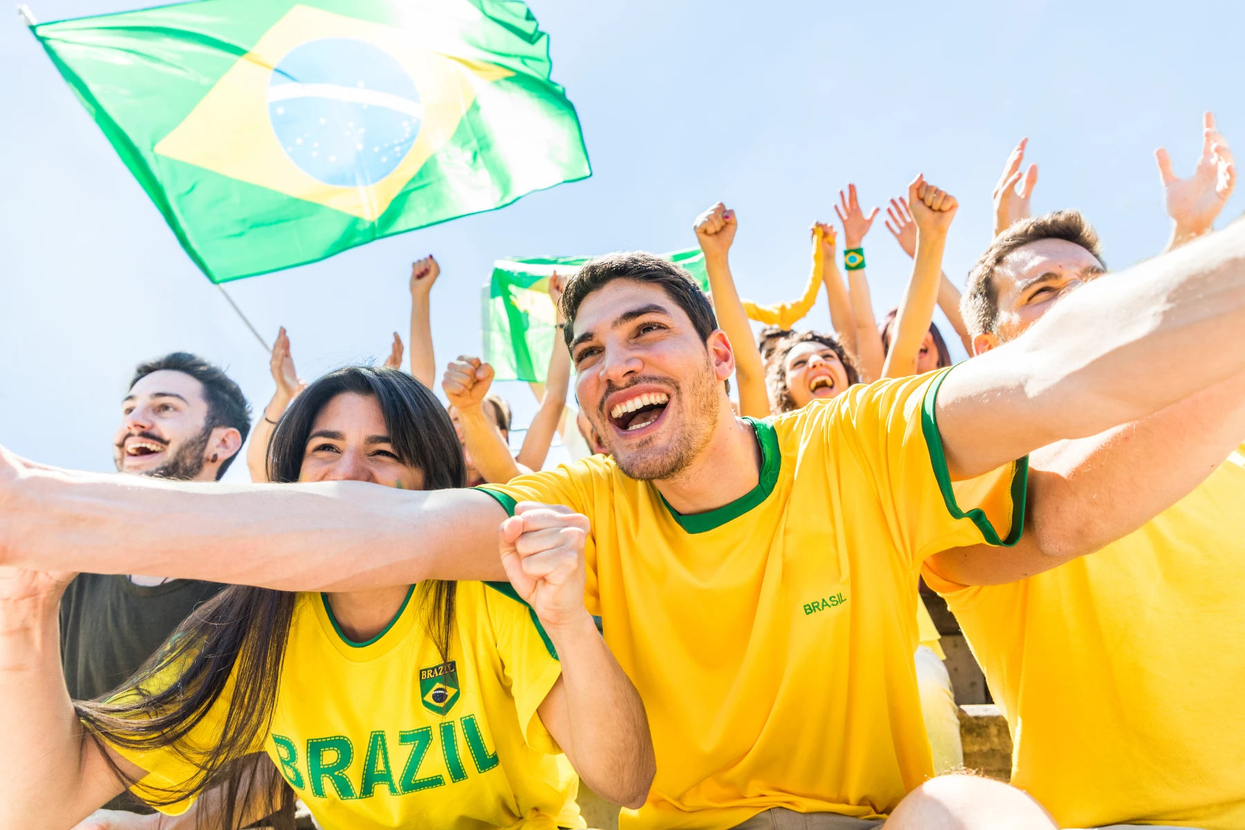 brazil-soccer-fans-celebrating-outdoors-with-flag-and-yellow-jerseys-hmg-plus. A group of Brazilian soccer fans in yellow and green Brasil jerseys celebrating outdoors under a blue sky, waving the Brazilian flag.