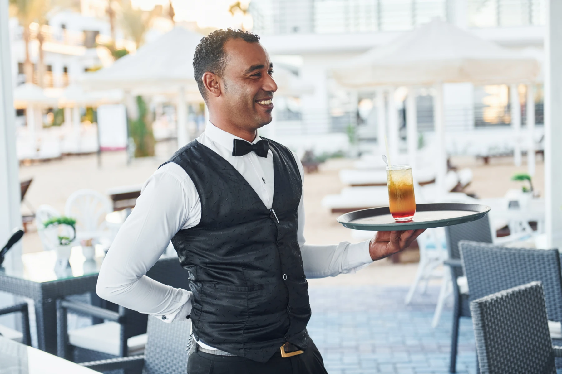 smiling-waiter-carrying-cocktail-tray-at-outdoor-resort-restaurant-hmg-plus. A smiling waiter in a black vest and bow tie carrying a tray with a layered cocktail at an outdoor resort restaurant.