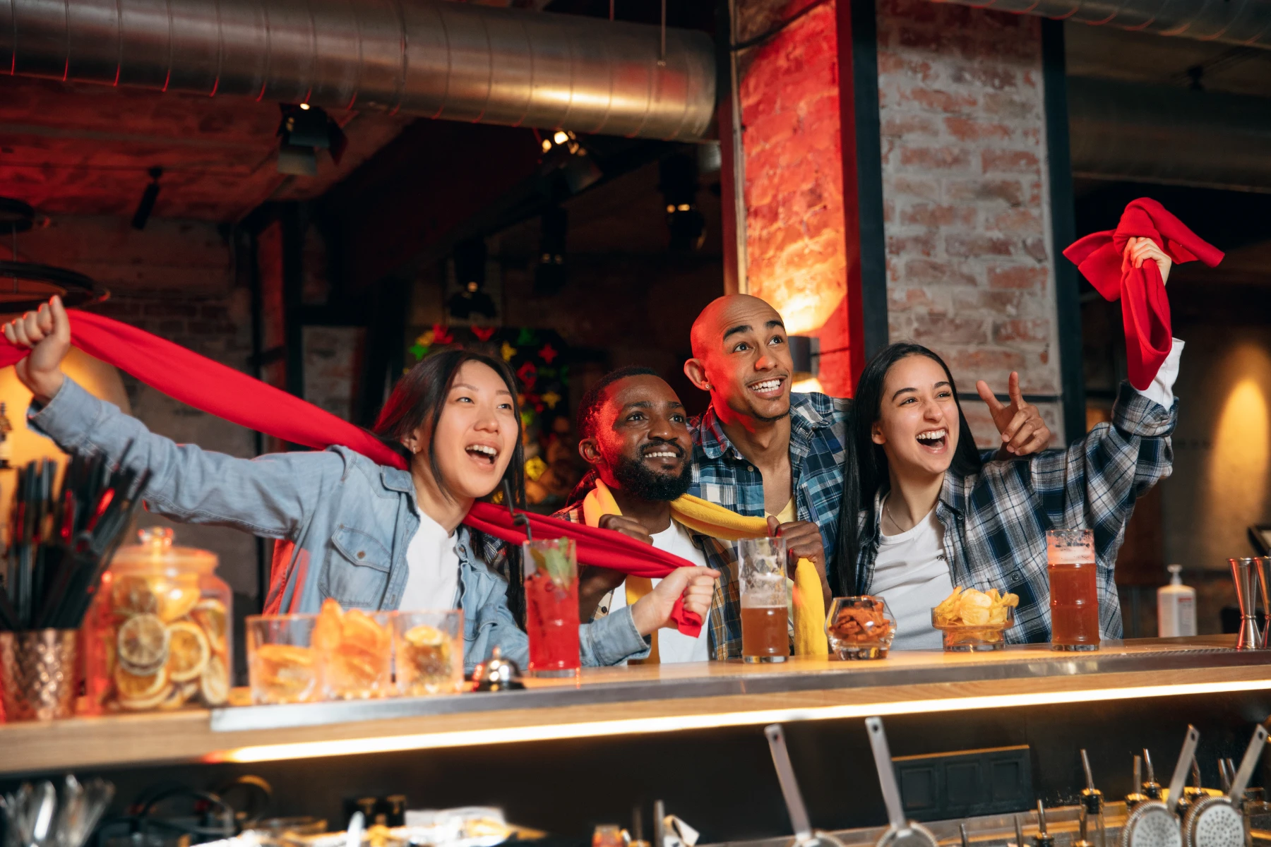 diverse-friend-group-cheering-at-sports-bar-with-red-scarves-hmg-plus. A diverse group of four friends cheering and waving red scarves at a sports bar with drinks and snacks on the counter.