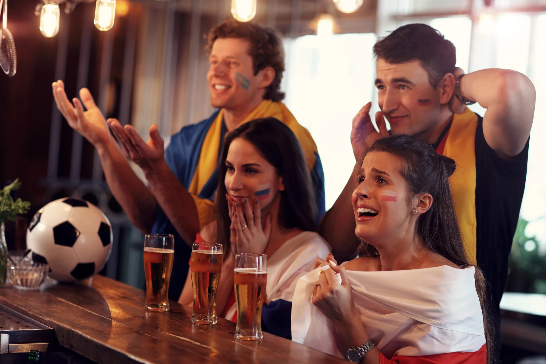 soccer-fans-watching-game-at-bar-with-face-paint-and-beer-hmg-plus. Four excited soccer fans with face paint watching a game at a bar, with beer glasses and a soccer ball on the counter.