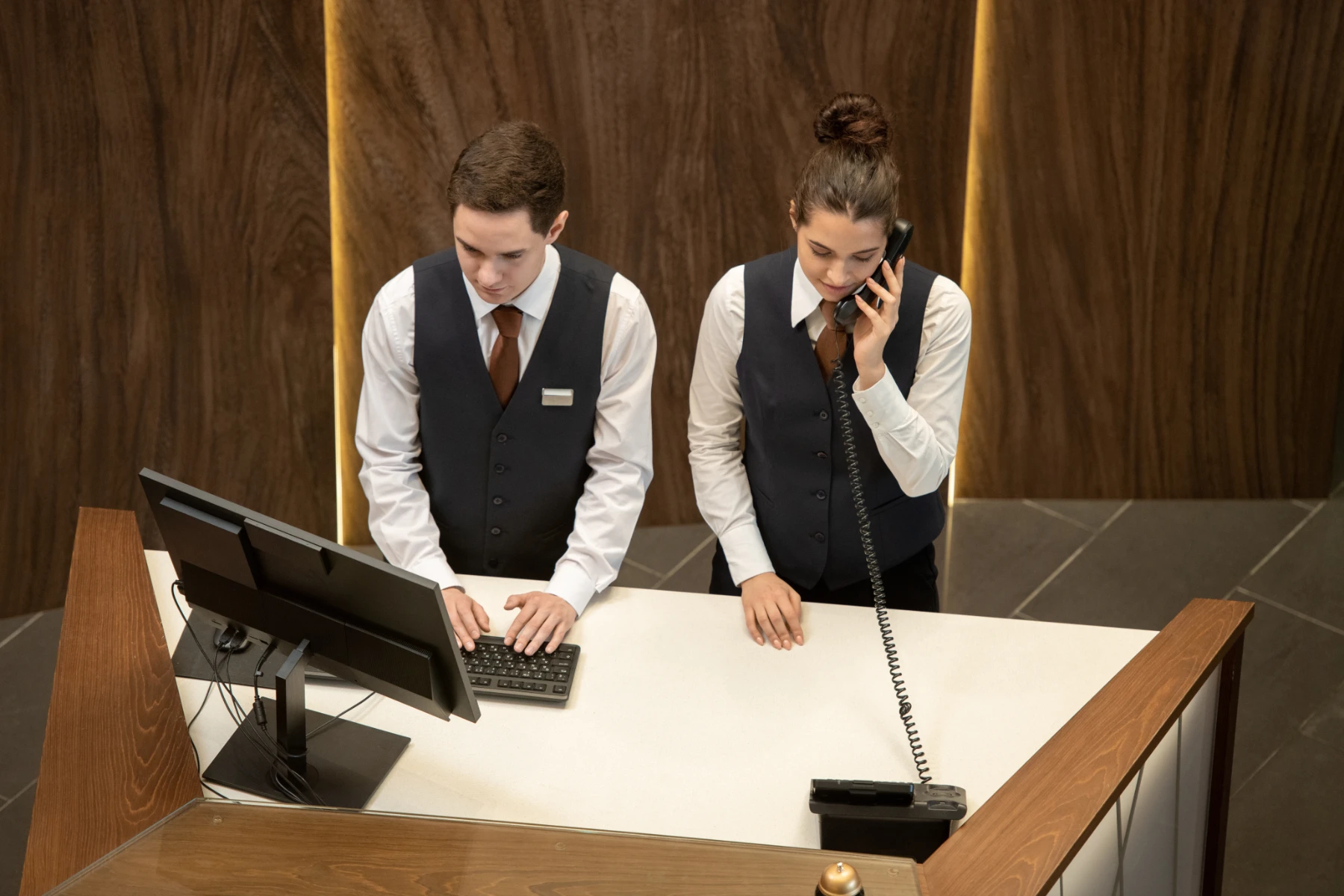hotel-front-desk-staff-working-at-reception-counter-hmg-plus. Two hotel front desk employees in matching navy vests working at a reception counter, one typing on a computer and one speaking on the phone.