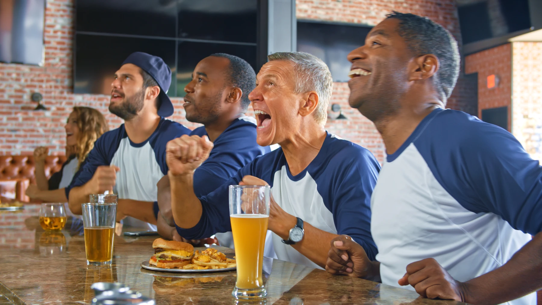 male-sports-fans-cheering-at-bar-in-matching-team-jerseys-hmg-plus. Four male sports fans in matching navy and white baseball-style jerseys cheering enthusiastically at a sports bar with beer and food.