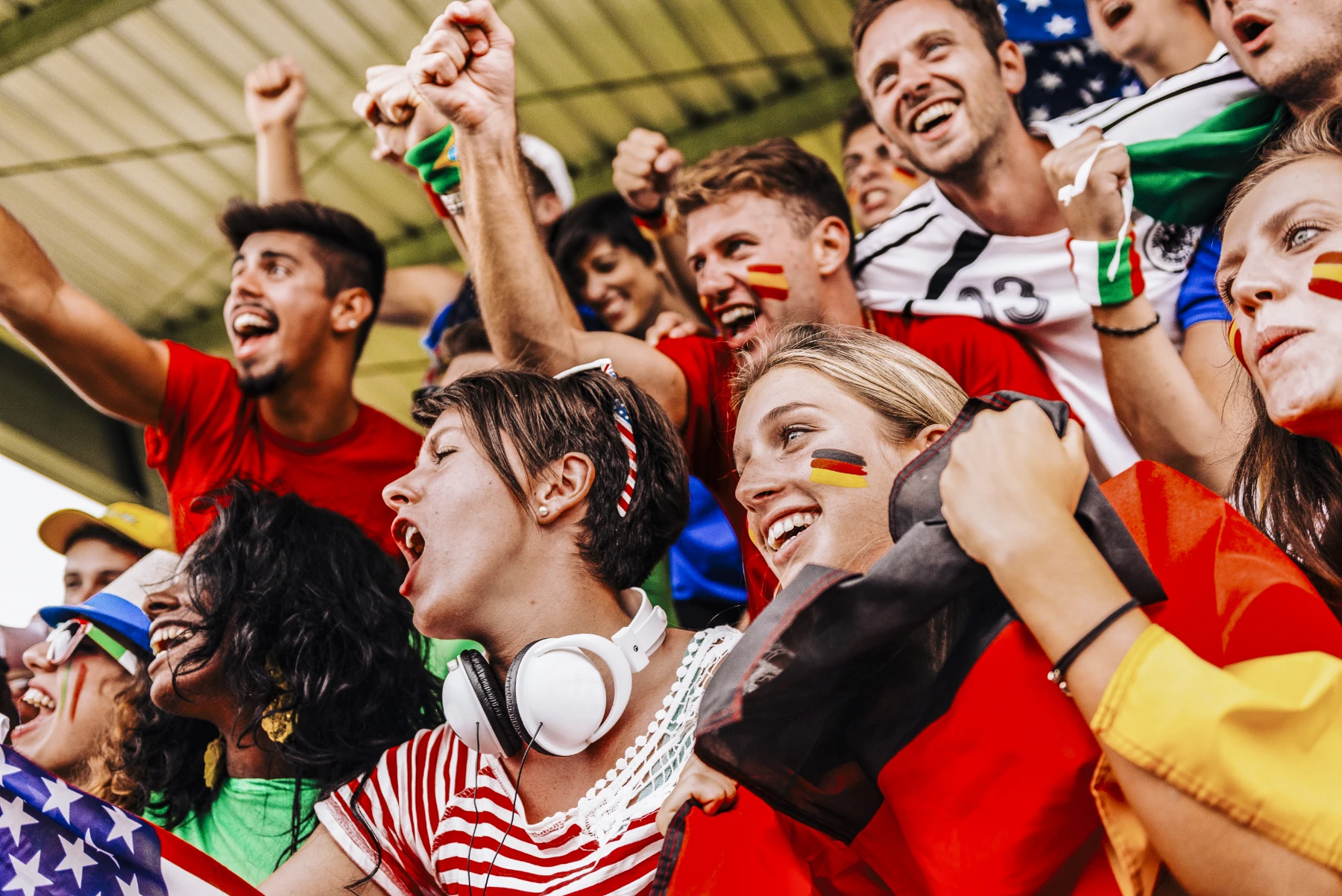 international-soccer-fans-celebrating-in-stadium-stands-hmg-plus. A diverse crowd of international soccer fans celebrating in stadium stands, with face paint and national flags from multiple countries.