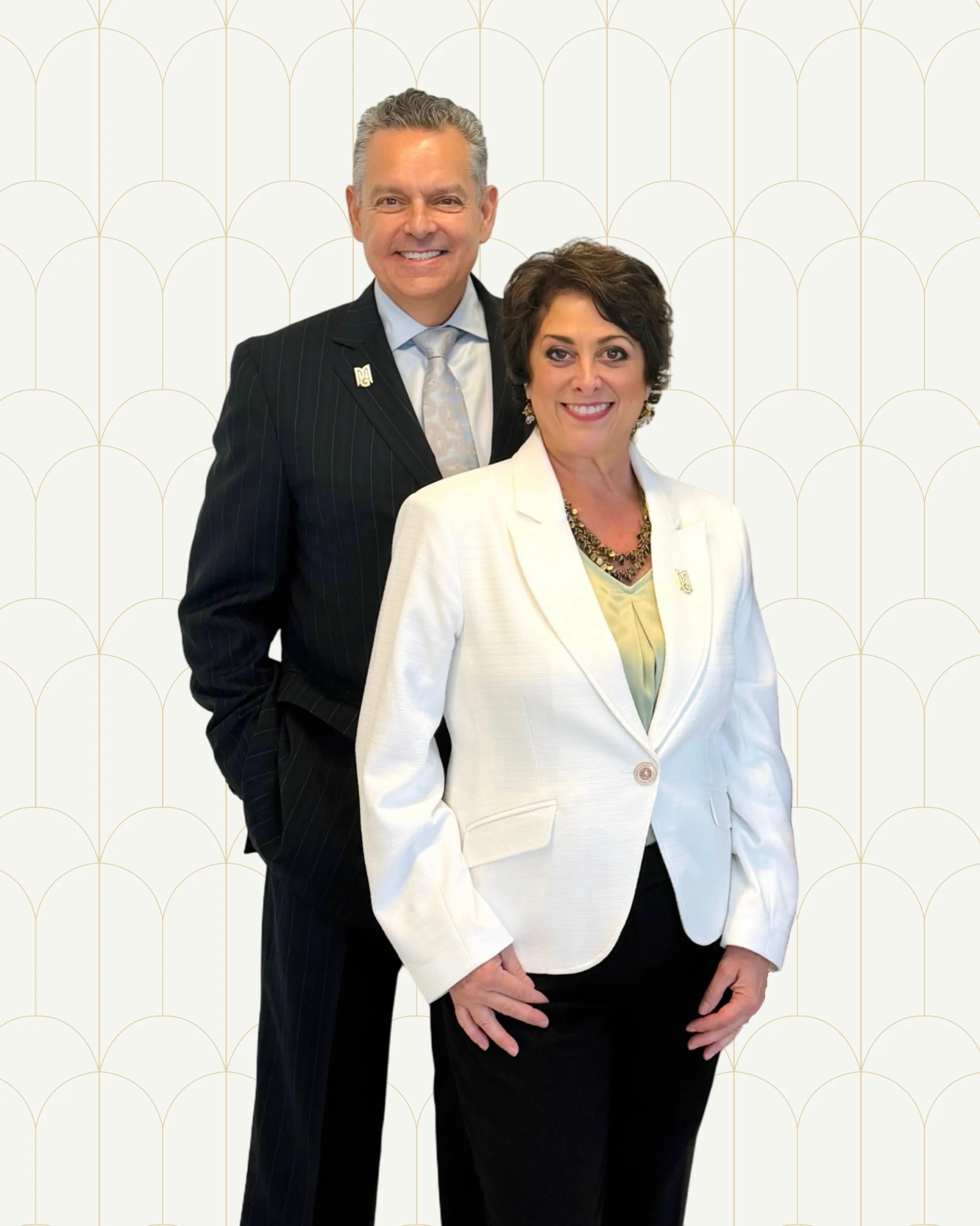 karen-and-michael-diperi-posing-for-formal-corporate-portrait A man in a dark pinstripe suit and a woman in a white blazer smile confidently together in a formal corporate portrait against a cream background with a gold arch pattern.